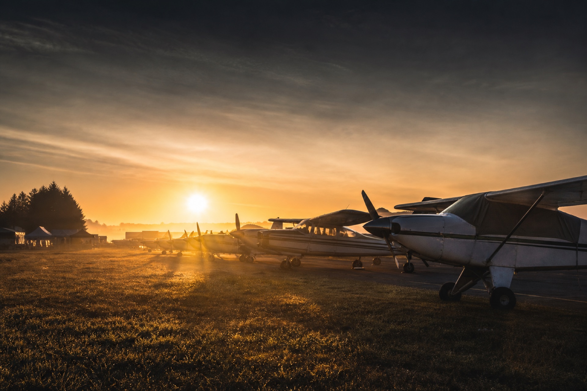 Line of parked aircraft at a fly-in, golden sunrise flaring through morning haze
